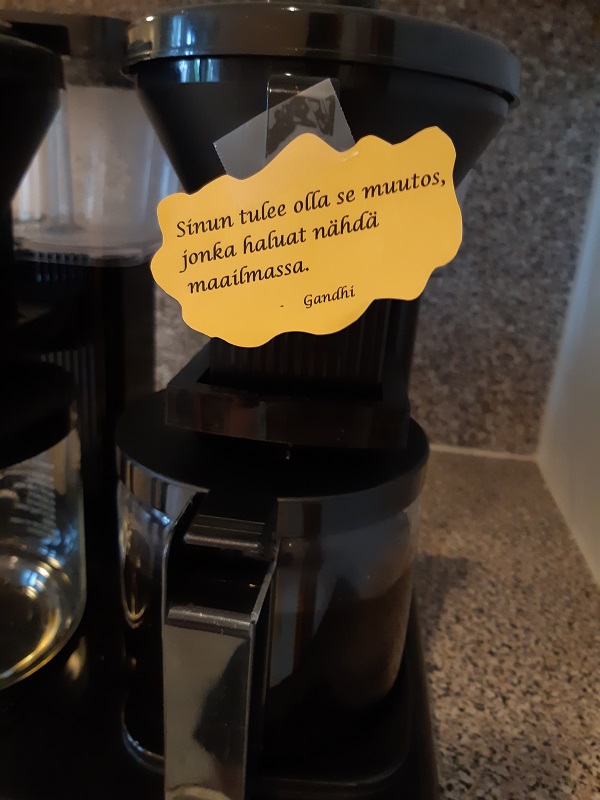 Kahvinkeittimeen kiinnitetty lappu, jossa lukee "Sinun tulee olla se muutos, jonka haluat nähdä maailmassa. -Ganhdi".