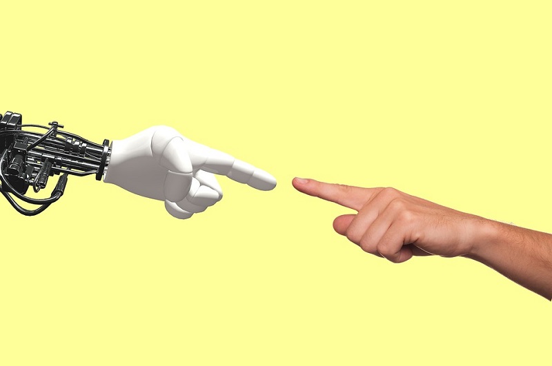 Robottikäden ja ihmiskäden sormet kohtaavat keltaista taustaa vasten.