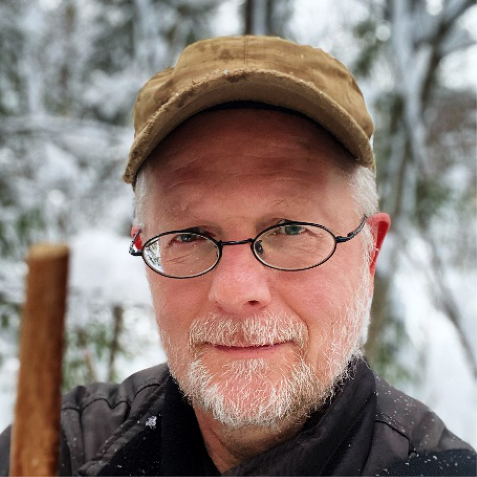 Porträttbild på man i glasögon och skärmmössa som står i snöig skog.